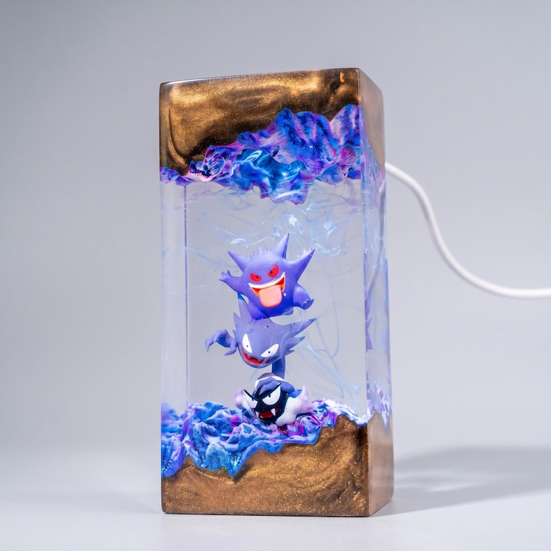 May include: A rectangular resin lamp featuring a clear centre section with three stacked purple Gengar-like figures. The top and bottom sections are made of a metallic bronze material with swirling blue and purple resin accents throughout.