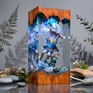 May include: A rectangular lamp made of wood and clear resin featuring a miniature figure of a character in a blue tunic holding a wooden structure above their head. The scene includes a rocky base and swirling white smoke effects inside the resin.