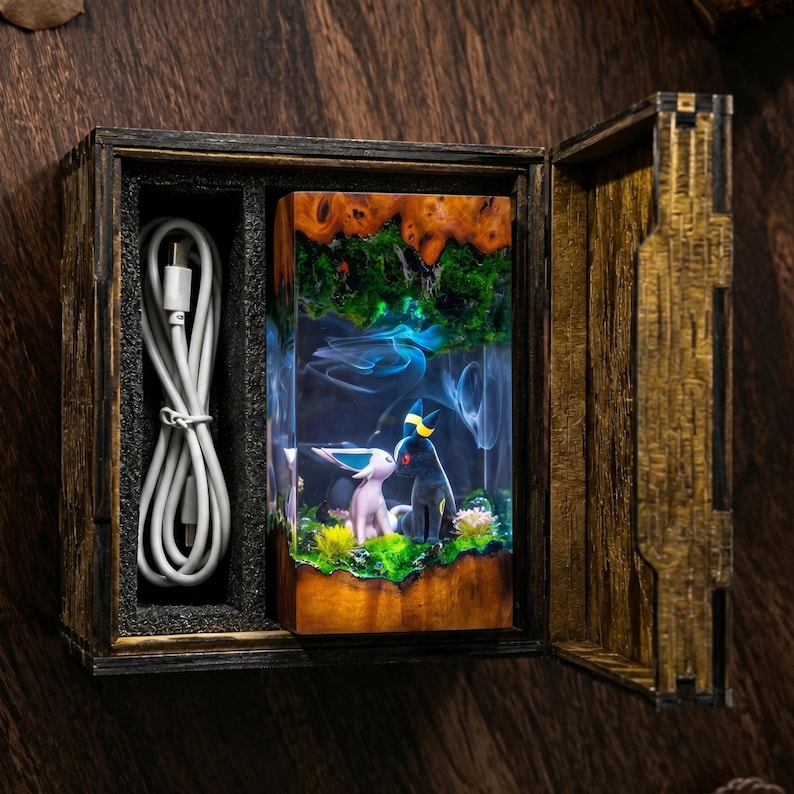 Op de afbeelding: Een rechthoekige houten doos met een verlicht harsdiorama met twee kleine dierfiguren, &eacute;&eacute;n roze en &eacute;&eacute;n zwart met gele accenten, omringd door groen mos en bloemen, vergezeld van een witte USB-oplaadkabel.