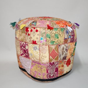 May include: A round, patchwork pouf with a beige base and a colorful, embroidered top. The patchwork design features various fabrics in shades of gold, red, purple, and orange. Small tassels adorn the sides. This decorative ottoman adds a bohemian touch to any living space.