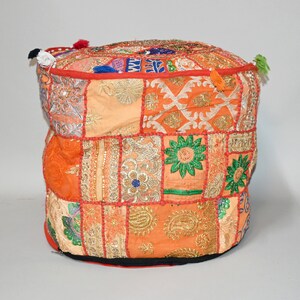 May include: A patchwork pouf in shades of orange, peach, and gold. The square panels are stitched together with red thread and adorned with embroidered floral and geometric patterns. Tassels in various colors are attached to the top edges.