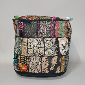 May include: A square, patchwork pouf with a black base and top. The sides are composed of various fabric squares in black, gold, pink, and green, adorned with intricate embroidery and embellishments. Small tassels in various colors decorate the top edge.