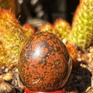 May include: A polished, spherical gemstone with a speckled pattern of red and brown hues, displayed on a small, red wooden stand. The background features spiky cacti with orange and green tones, creating a natural, earthy aesthetic.