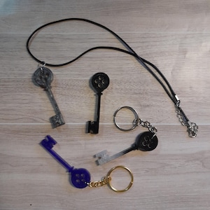 Pode incluir: Três pingentes em forma de chave com um design circular na parte superior. Um pingente é prateado, um é preto e um é azul. Os pingentes prateado e preto estão em uma corrente prateada, e o pingente azul está em uma corrente dourada.
