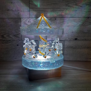 Puede incluir: Un carrusel de vidrio transparente con una base de purpurina dorada y una luz blanca en el interior. El carrusel tiene tres caballos y un techo de purpurina dorada.