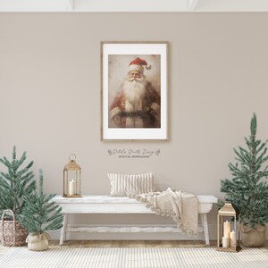 May include: A framed painting of Santa Claus with a red hat and white beard. The painting is hanging on a wall with a white bench in front of it. There are two small artificial Christmas trees on either side of the bench.