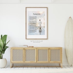 May include: A framed print with a poem titled "Footprints" about walking through life's challenges. The poem is printed on a white background with a watercolor-style image of a person walking on a beach with their footprints in the water. The image is framed in a light wood frame.