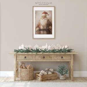 May include: A rustic wooden console table with three drawers, decorated with a garland of pine branches and white candles. There are small white houses and a small potted Christmas tree on the table. A framed print of a vintage Santa Claus illustration hangs on the wall above the table.