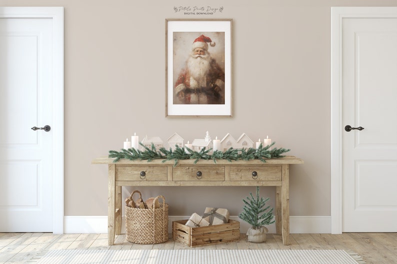 May include: A rustic wooden console table with three drawers, decorated with a garland of pine branches and white candles. There are small white houses and a small potted Christmas tree on the table. A framed print of a vintage Santa Claus illustration hangs on the wall above the table.