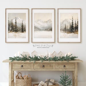 May include: Three framed watercolor paintings of snowy mountain landscapes with evergreen trees. The paintings are displayed on a white wall above a wooden console table with a Christmas garland and decorative houses.