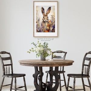 Puede incluir: Una pintura de acuarela de un conejo marr&oacute;n con orejas largas en un campo de flores silvestres p&uacute;rpuras y amarillas. La pintura est&aacute; enmarcada en un marco de madera simple y est&aacute; colgada en una pared blanca.