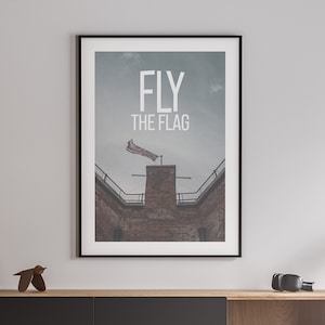 Puede incluir: Una impresión enmarcada con un fondo gris y el texto "FLY THE FLAG" en blanco. La imagen muestra un edificio de ladrillo con una bandera ondeando al viento.
