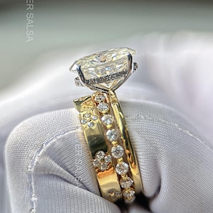 May include: A three-piece ring set featuring a large, cushion-cut diamond engagement ring with a halo setting, and two gold bands with smaller diamonds. The rings are set against a white background.