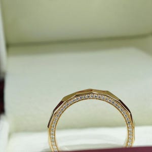 May include: A gold ring with a geometric design, set with small, clear gemstones. The ring is displayed in a burgundy jewelry box with the text "SILVER SALSA STUDIO". The ring's band has a faceted appearance.