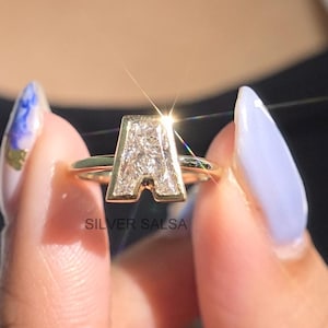 Pode incluir: Um anel dourado com a letra "A" incrustada com pequenas pedras transparentes. O anel é segurado, mostrando seu design. O nome da marca "SILVER SALSA" é visível na parte inferior do anel.