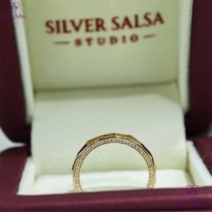 May include: A gold ring with a geometric design, set with small, sparkling diamonds. The ring is displayed inside a burgundy jewelry box with the text "Silver Salsa Studio".