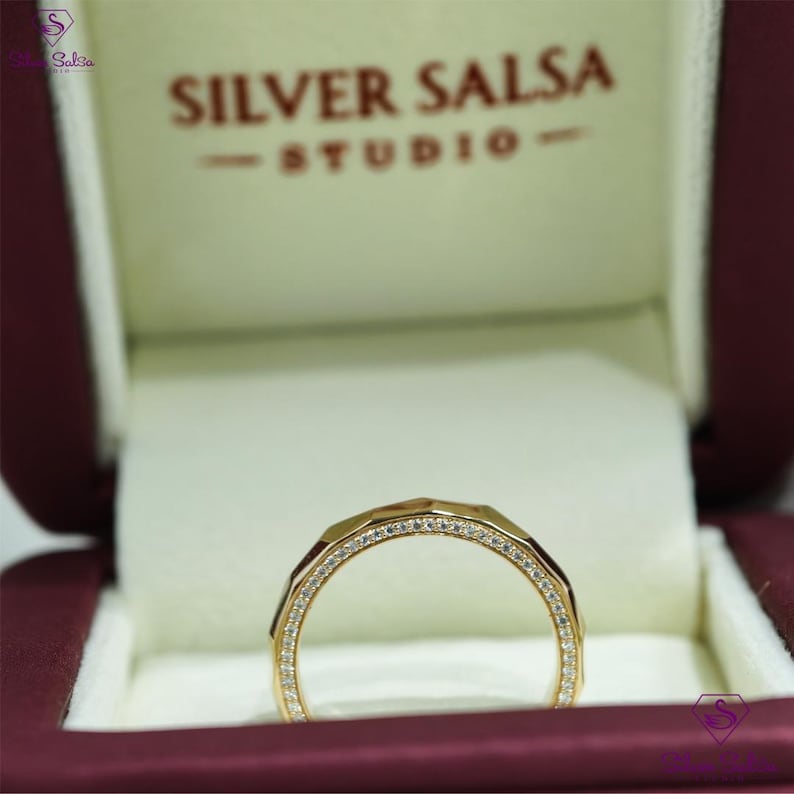 May include: A gold ring with a geometric design, set with small, sparkling diamonds. The ring is displayed inside a burgundy jewelry box with the text "Silver Salsa Studio".