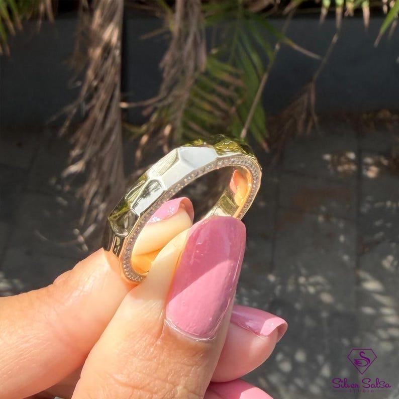 May include: A gold ring with a geometric design and a band of small, clear stones. The ring has a white enamel section and is held up by a person with pink nail polish.