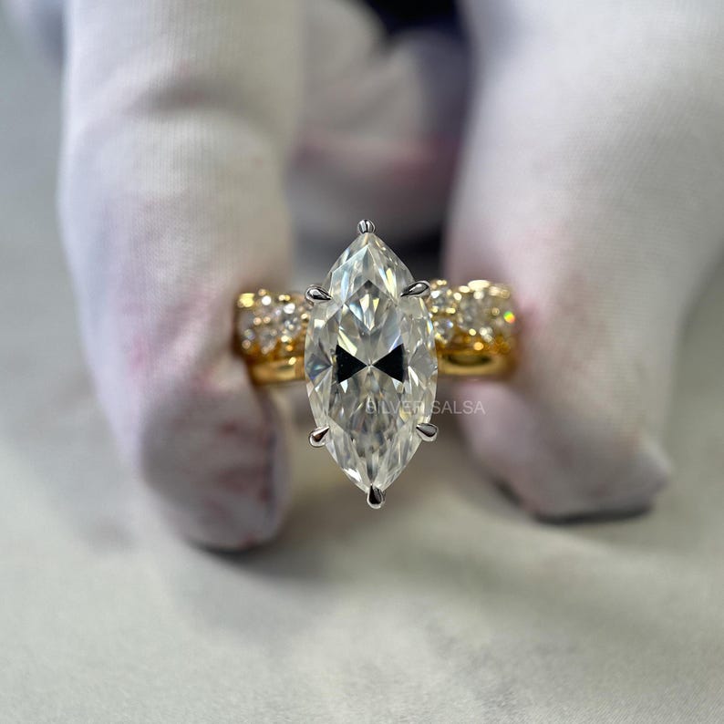 May include: A marquise-cut diamond ring with a gold band. The ring features a large, clear, marquise-cut diamond as the centerpiece, with smaller diamonds set along the band. The ring is held by a gloved hand.