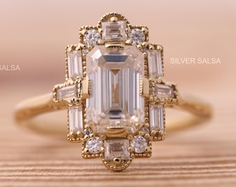 May include: A gold-toned ring featuring an emerald-cut center stone, surrounded by smaller baguette and round-cut stones. The ring has an Art Deco design with intricate detailing and the text "SILVER SALSA".