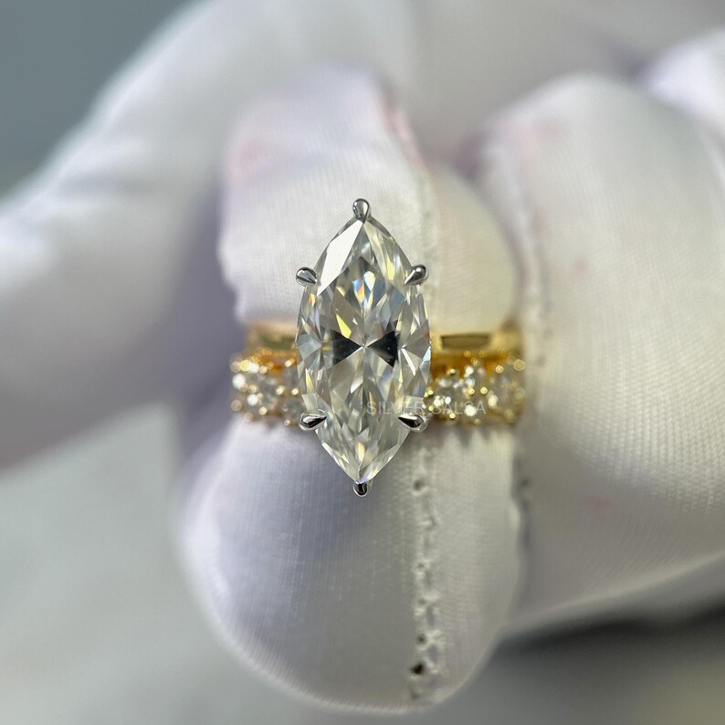 May include: A marquise-cut diamond engagement ring with a gold band, set on a white background. The diamond is the focal point, with a clear, brilliant cut. A second gold band with smaller stones is visible.