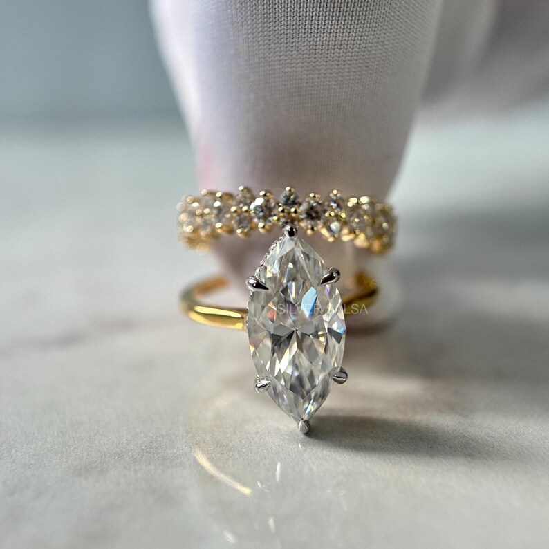 May include: A marquise-cut diamond engagement ring with a gold band, set against a blurred background. A second gold band with small diamonds is visible behind the engagement ring. The rings are displayed on a white surface.