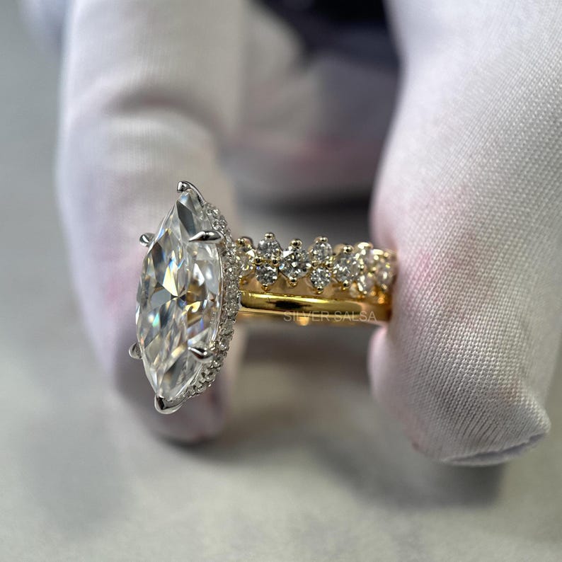 May include: A marquise-cut diamond engagement ring with a halo of smaller diamonds. The ring has a gold band and is held by a gloved hand. A second gold band with small diamonds is also visible.