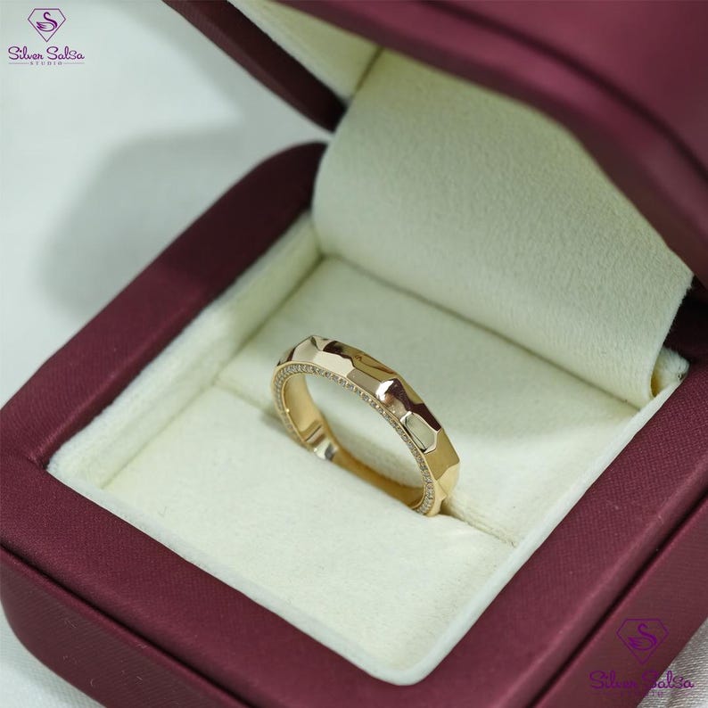 May include: A gold ring with a geometric design, set in a burgundy jewelry box. The ring features a series of facets and a row of small stones along the edge. The jewelry box has a white interior.