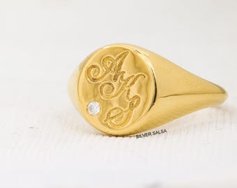 May include: A gold signet ring with a round face featuring the intertwined letters "A" and "G" and a small diamond accent. The ring is highly polished and has a smooth, rounded band. The text "SILVER SALSA" is visible.
