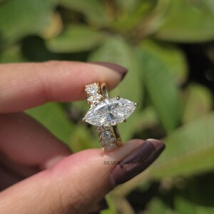 May include: A gold-colored engagement ring set featuring a marquise-cut diamond and a band with smaller accent stones. The ring is held up against a blurred green background, showcasing the sparkle of the gemstones.