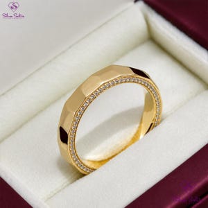 May include: A gold ring with a geometric design and a row of small, clear stones. The ring is displayed in a white box with a burgundy interior, showcasing its polished finish and intricate details.