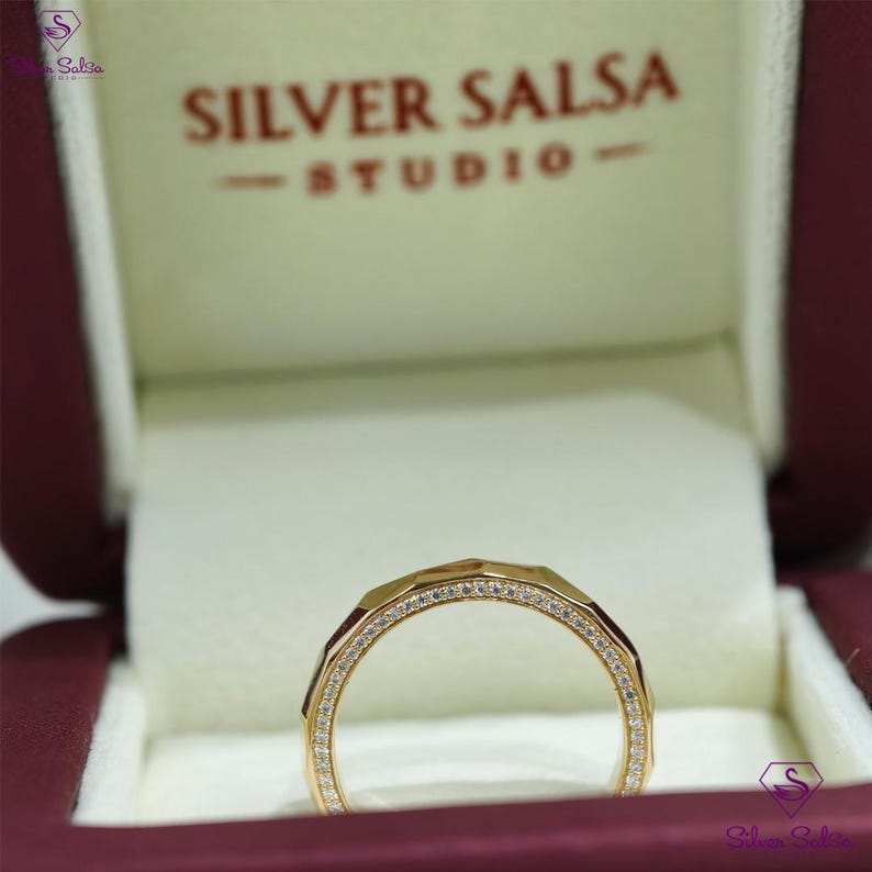 May include: A gold ring with a geometric design, set with small, clear gemstones. The ring is displayed in a burgundy jewelry box with the text "SILVER SALSA STUDIO". The ring's band has a faceted appearance.