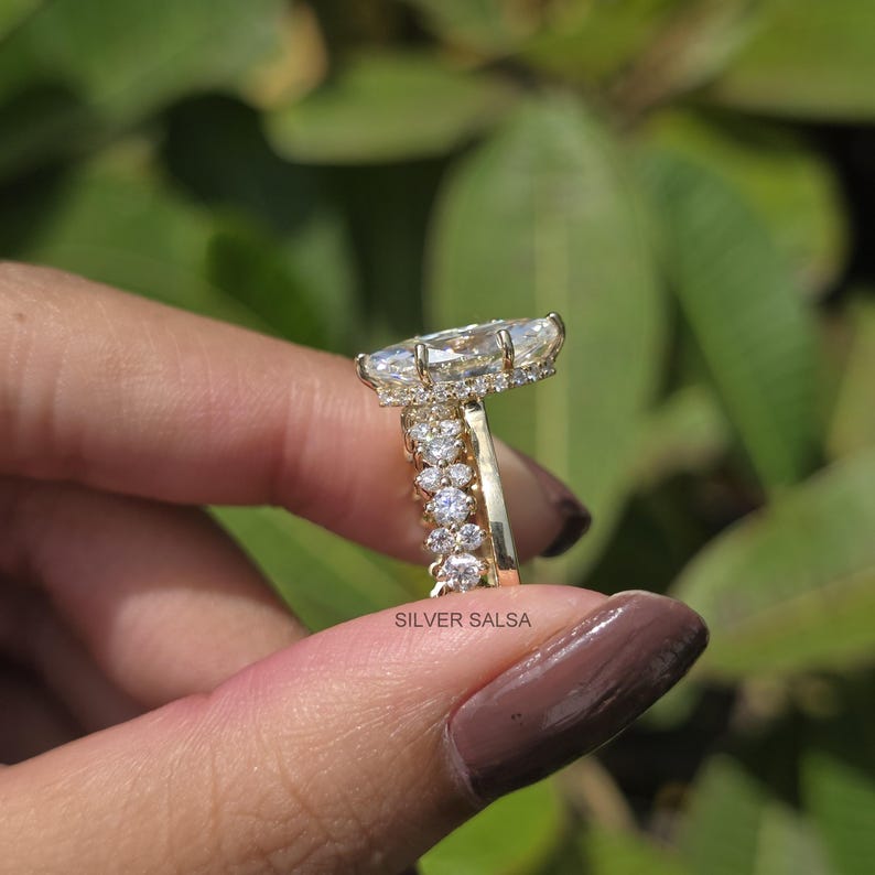 May include: A gold engagement ring with a large, pear-shaped diamond center stone. The ring features a halo of smaller diamonds around the center stone and a band with additional diamonds. The ring is held up against a green background.