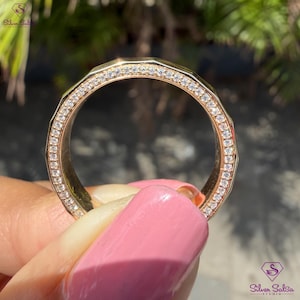 May include: A gold-colored ring with a faceted design and a row of small, clear gemstones set around the outer edge. The ring is held up by a person with pink nail polish.
