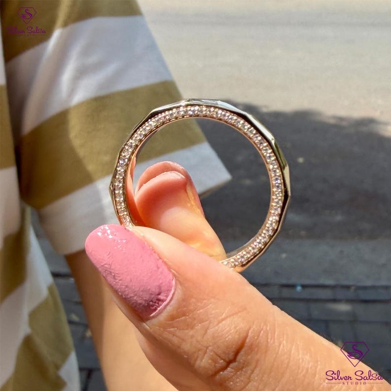 May include: A gold-toned ring with a geometric design and encrusted with small, clear gemstones. The ring is held up, showcasing its intricate details and sparkling appearance. The ring is a piece of jewelry.