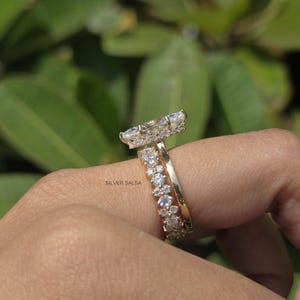 May include: A gold ring set with a large, marquise-cut diamond and a smaller band with multiple round diamonds. The rings are on a person's finger, with a blurred green background.