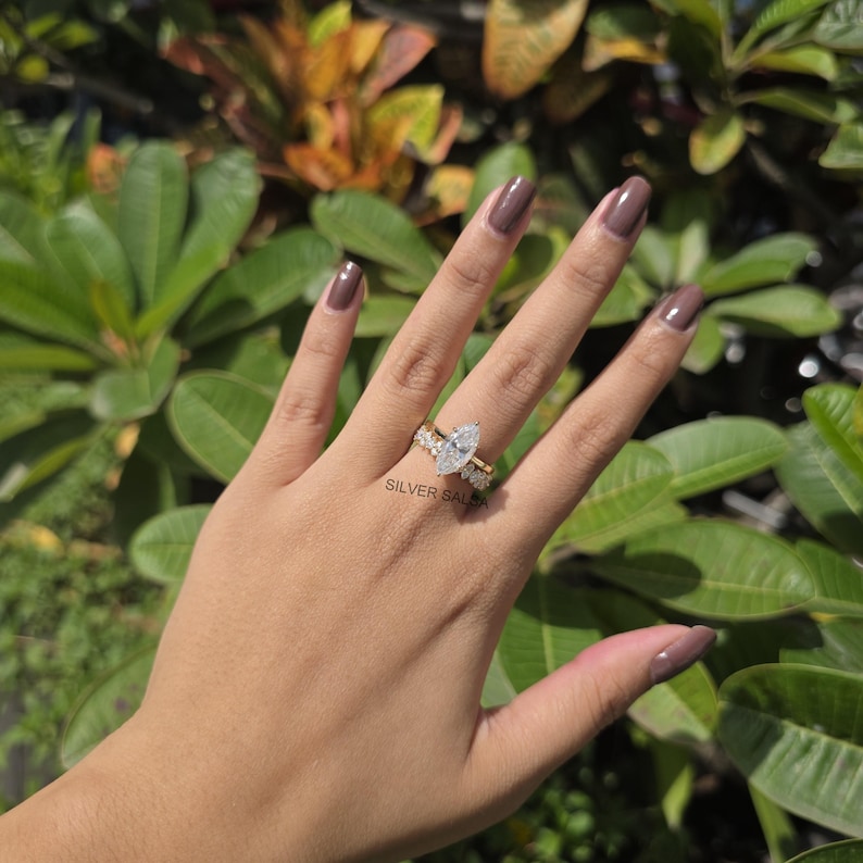 May include: A gold ring with a large pear-shaped diamond center stone and smaller diamonds on the band. The ring is on a hand with brown nail polish, set against a backdrop of green foliage. The text "SILVER SALSA" is visible.