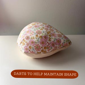 May include: A floral print newsboy cap with a pink, yellow, and green pattern on a white background. The cap has a beige brim and the text "DARTS TO HELP MAINTAIN SHAPE" in a brown banner.