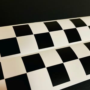 May include: Two rolls of checkered pattern vinyl. The pattern consists of alternating black and white squares. The rolls are slightly curved, with the pattern visible against a dark background. The squares are uniform in size.