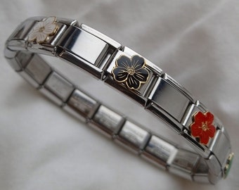 Op de afbeelding: Een roestvrijstalen armband met rechthoekige schakels. De armband heeft bloemvormige bedels in wit, zwart en rood met gouden accenten. Gepresenteerd op een witte, getextureerde achtergrond.