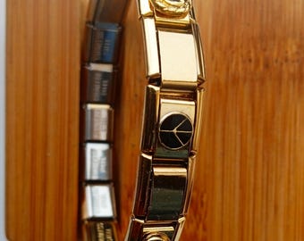 Op de afbeelding: Een gouden armband met bedels met een zwarte vredesduifbedel, een rode hartbedel en een zwarte en gouden bloemenbedel.