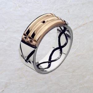 Op de afbeelding: Tweekleurige ring met een goudkleurige bovenkant en een zilverkleurige band. Het gouden gedeelte heeft een geribbeld ontwerp met een klein donker accent. De zilveren band heeft een geometrisch patroon en een oneindigheidssymbool.
