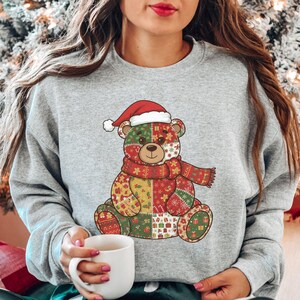 May include: A light gray sweatshirt featuring a colorful patchwork teddy bear design. The bear wears a Santa hat and a red scarf. The design incorporates red, green, and yellow patterns, perfect for the holiday season.