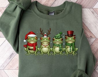 May include: An olive green sweatshirt featuring a festive graphic of four green frogs. Each frog is dressed in holiday attire, including a Santa hat, reindeer antlers, a green and white polka dot hat, and a top hat. The frogs are wearing scarves and are ready for the holidays.
