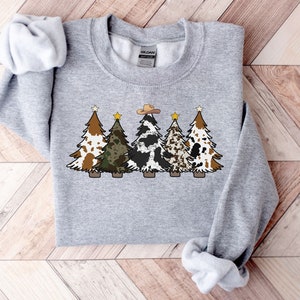 May include: A grey sweatshirt featuring a design of five Christmas trees. The trees are decorated with various patterns, including cowhide, camouflage, and brown and white spots. A cowboy hat sits atop the central tree.