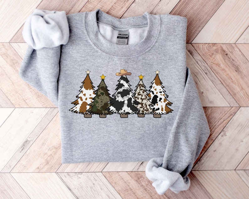 May include: A grey sweatshirt featuring a design of five Christmas trees. The trees are decorated with various patterns, including cowhide, camouflage, and brown and white spots. A cowboy hat sits atop the central tree.