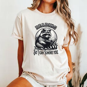 May include: A cream-colored t-shirt with a black and white illustration of a raccoon reading a book. The text "Reading so I can ignore you." is printed on the shirt.