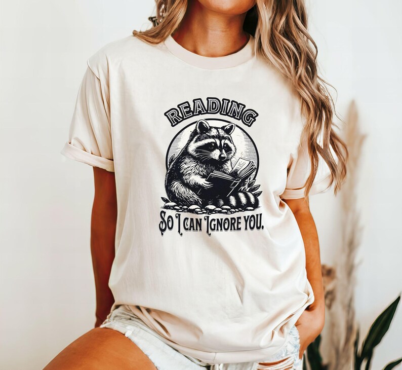 May include: A cream-colored t-shirt with a black and white illustration of a raccoon reading a book. The text "Reading so I can ignore you." is printed on the shirt.