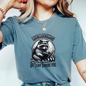 May include: A blue t-shirt with a graphic of a raccoon reading a book. The text on the shirt reads "Reading so I can ignore you."