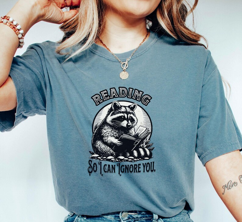 May include: A blue t-shirt with a graphic of a raccoon reading a book. The text on the shirt reads "Reading so I can ignore you."